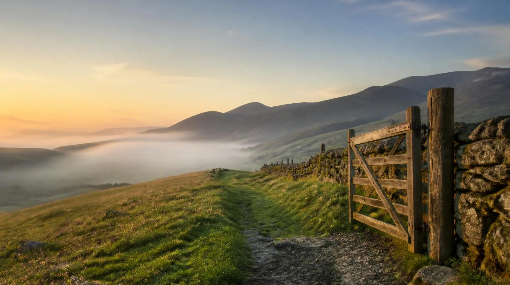 An open wooden gate on a grassy hill path at sunrise, with mist in the valley below